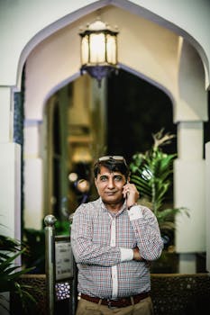 A South Asian man in casual attire talks on his mobile phone under a lit archway.